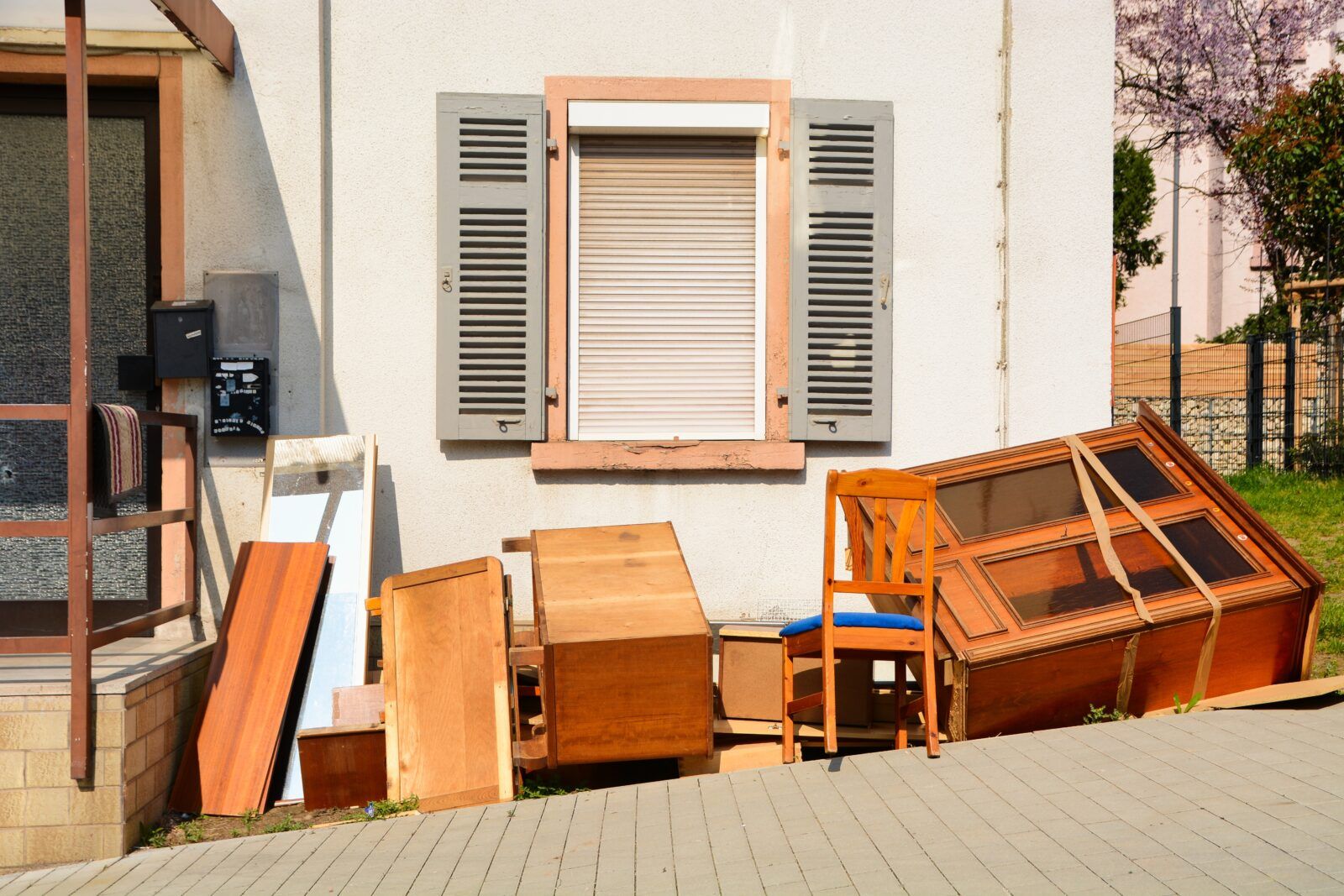 Pile of discarded wooden furniture in front of a light-colored building with a shuttered window.