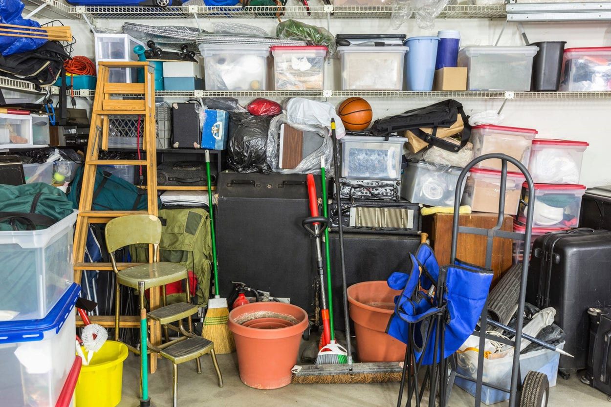 Cluttered garage with storage bins, tools, a ladder, and other items on shelves and floor.