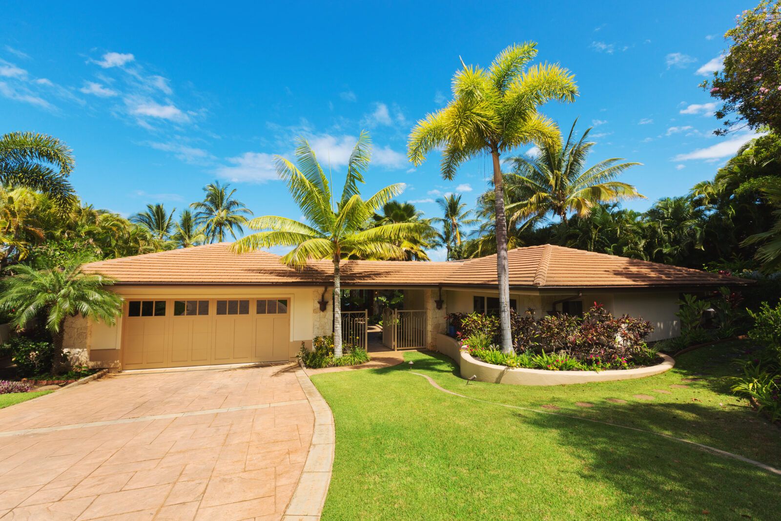House with beige exterior, tan roof, palm trees, and a concrete driveway under a blue sky.