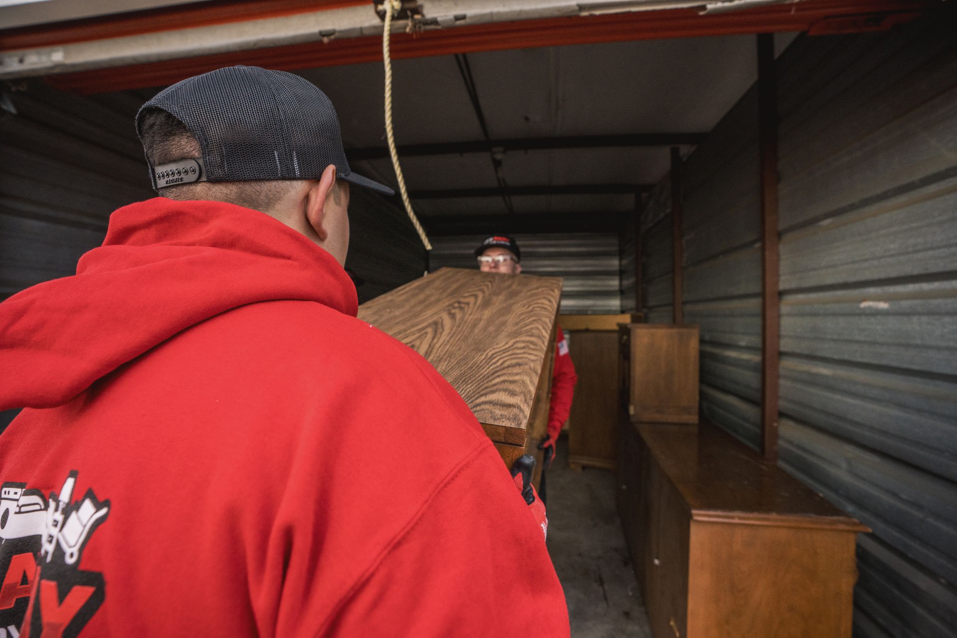 Man in red hoodie helping another carry wooden furniture into a storage unit.
