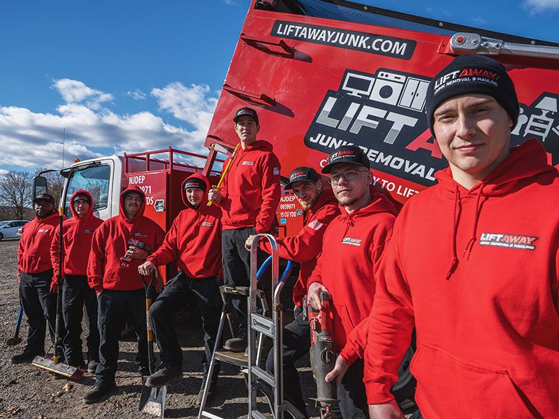 Team of junk removers in red hoodies stands next to a red truck on a sunny day.