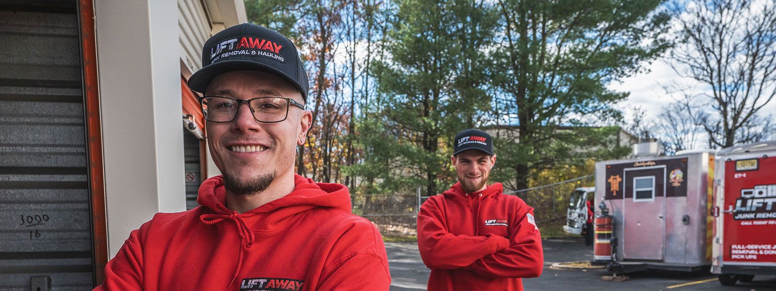 Two men in red hoodies and hats smiling, in front of trucks and storage units.
