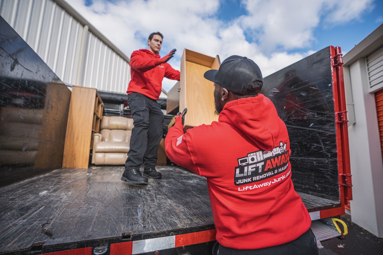 Two men in red hoodies load a wooden drawer into a truck bed under a cloudy sky.