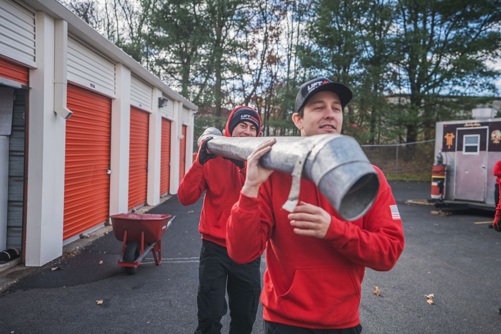 Two men in red hoodies carry large metal pipe in front of storage units.