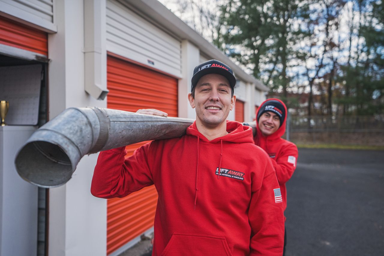 Two men in red hoodies smile, one carrying a large metal pipe, storage units in the background.