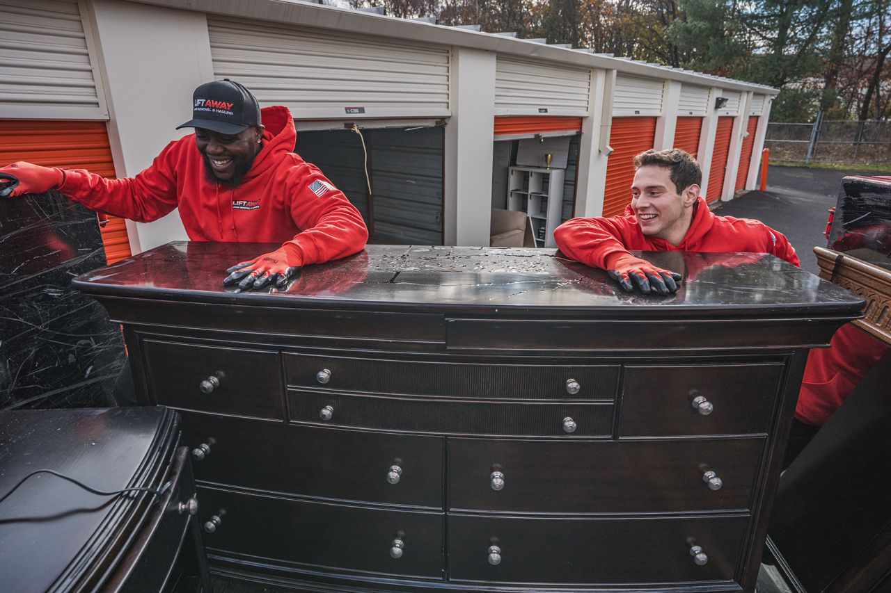 Two movers in red gear carrying a dark wood dresser, smiling, near storage units.