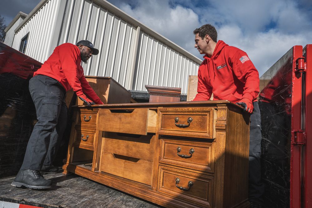 Two movers in red unloading a wooden dresser from a truck.
