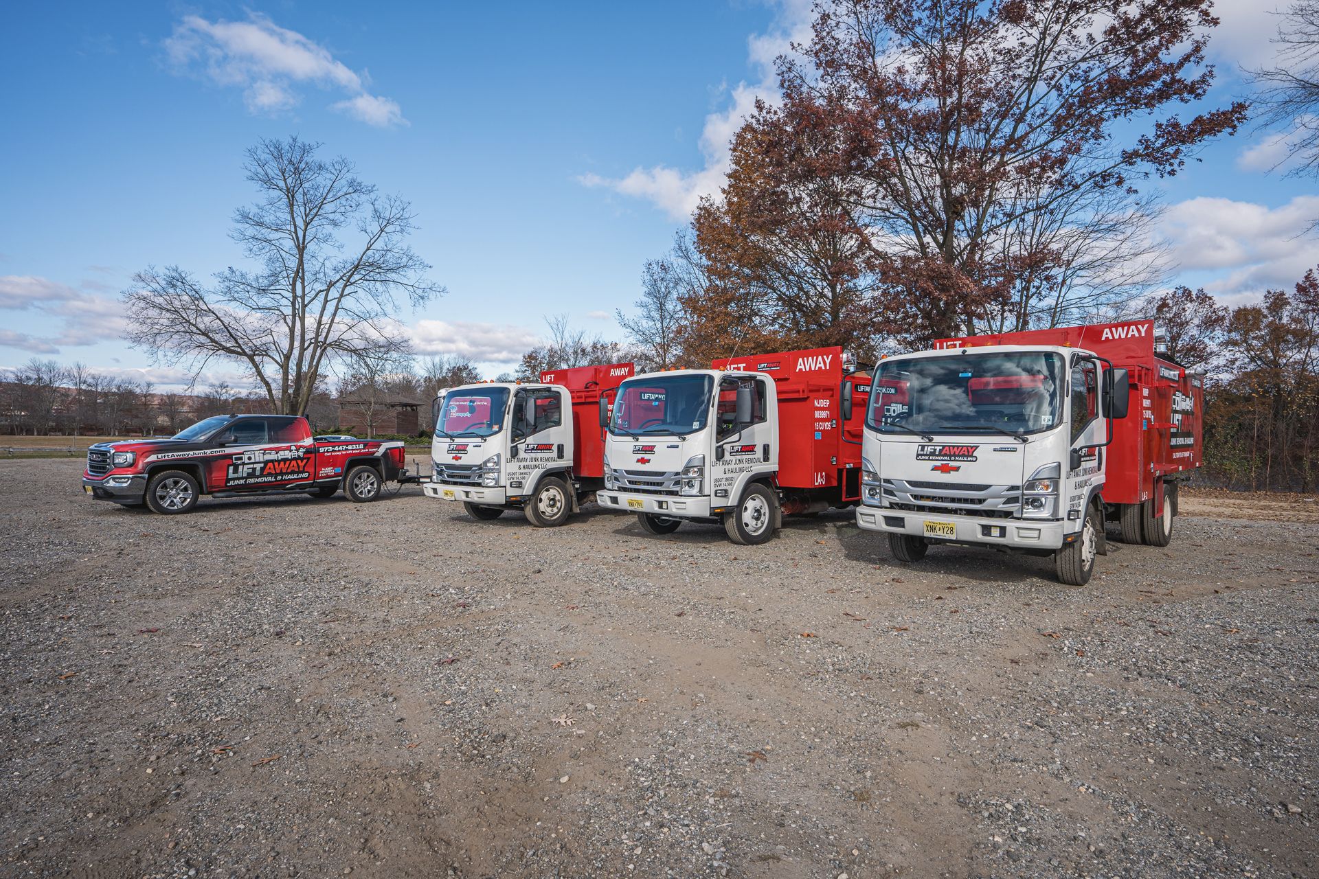 Four service trucks parked on gravel under a blue sky with fall foliage in the background.