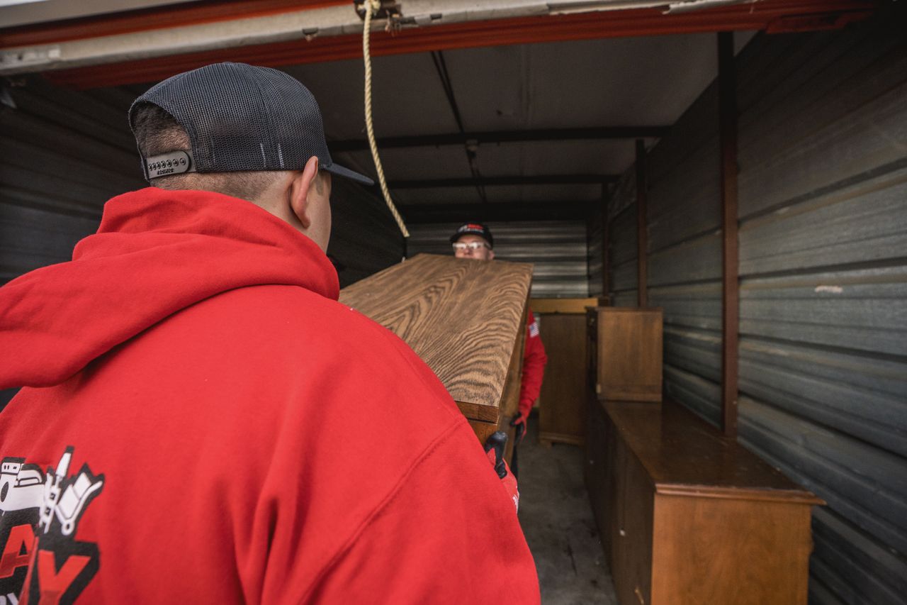 Two men carrying a wooden board into a storage unit, one in a red hoodie.