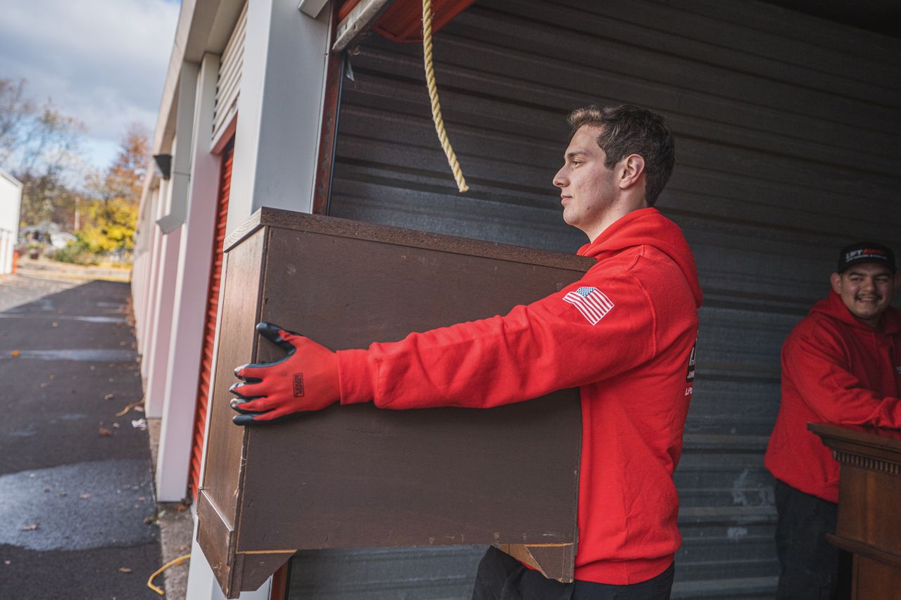 Two men in red hoodies carrying a brown cabinet out of a storage unit.