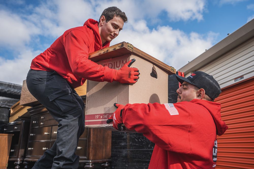Two movers in red hoodies lift a cardboard box, likely at a storage facility.