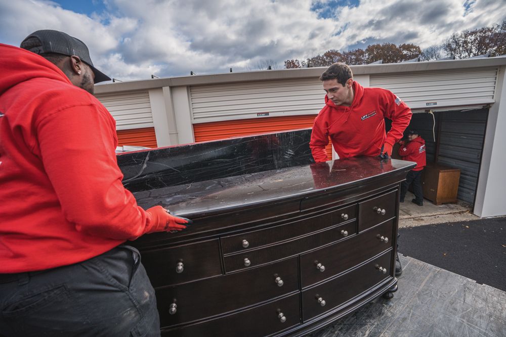 Movers in red uniforms loading a dark wood dresser into a storage unit, cloudy sky.