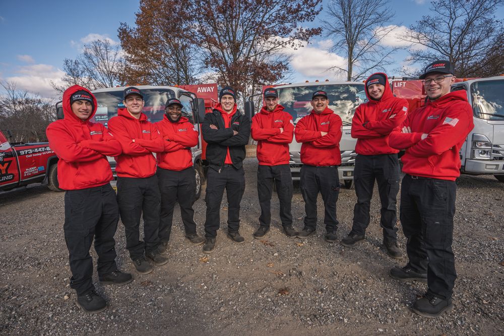 A group of eight tow truck operators in red jackets stand in front of their trucks.