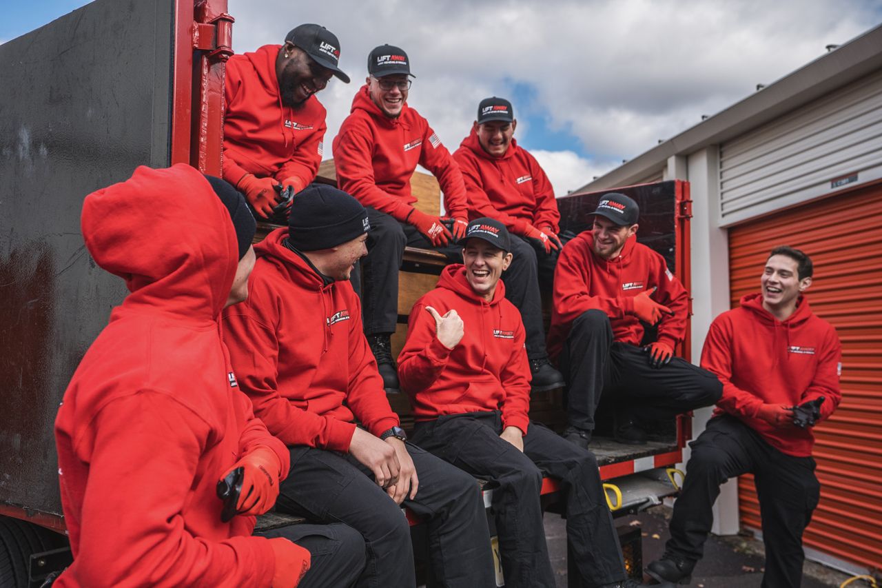 A group of movers in red hoodies and black pants are smiling while on the back of a truck.