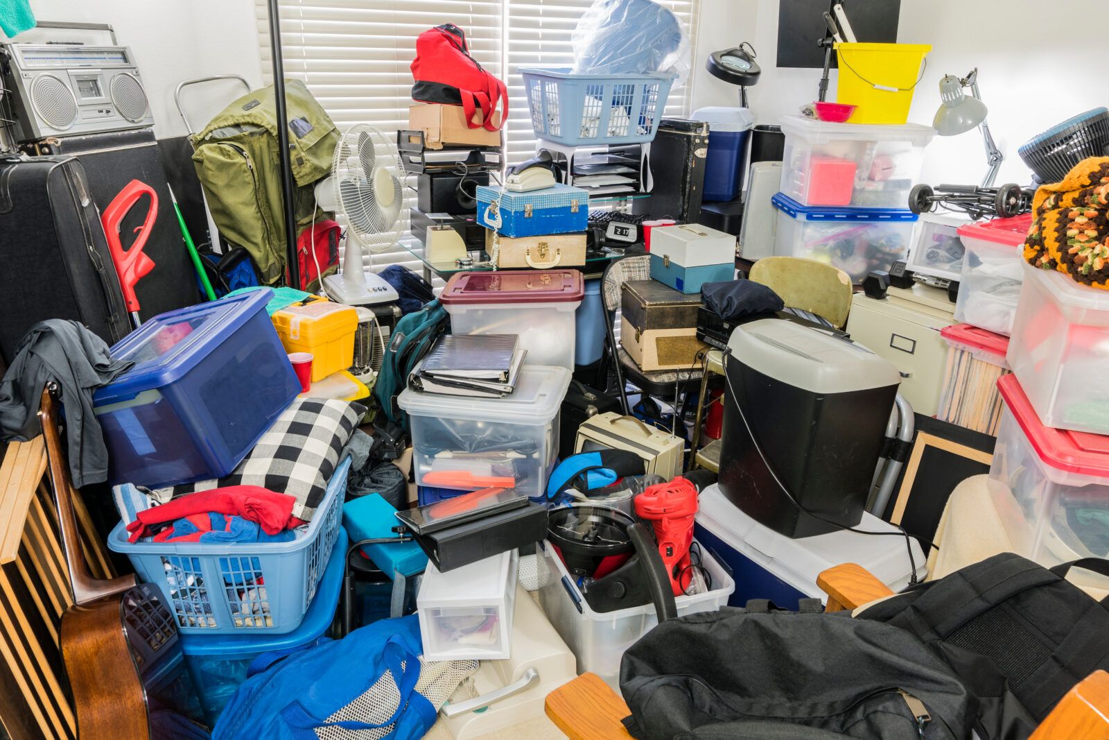 Cluttered room with boxes, clothes, and items piled up against a wall with a closed window.