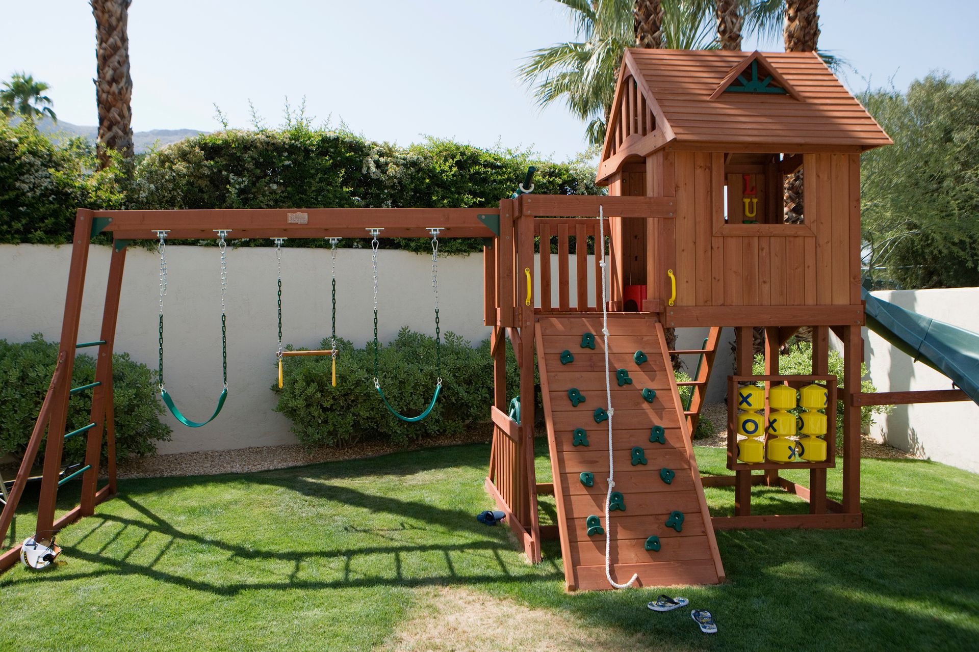 Wooden playset with swings, climbing wall, and slide on green lawn.