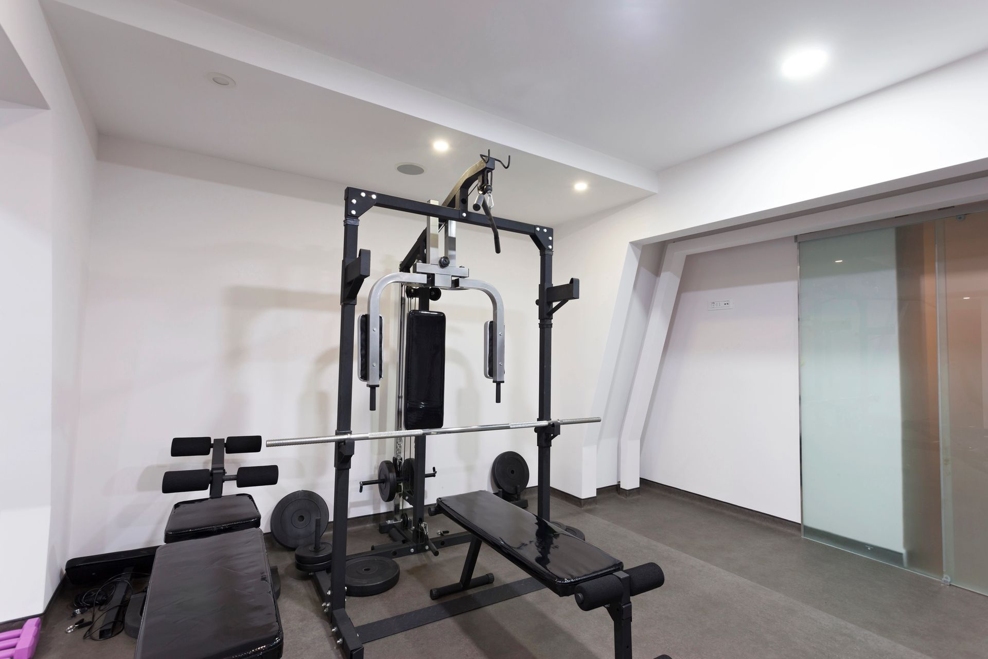 Gym with weight bench, multi-station machine, and weights. White walls, dark flooring.