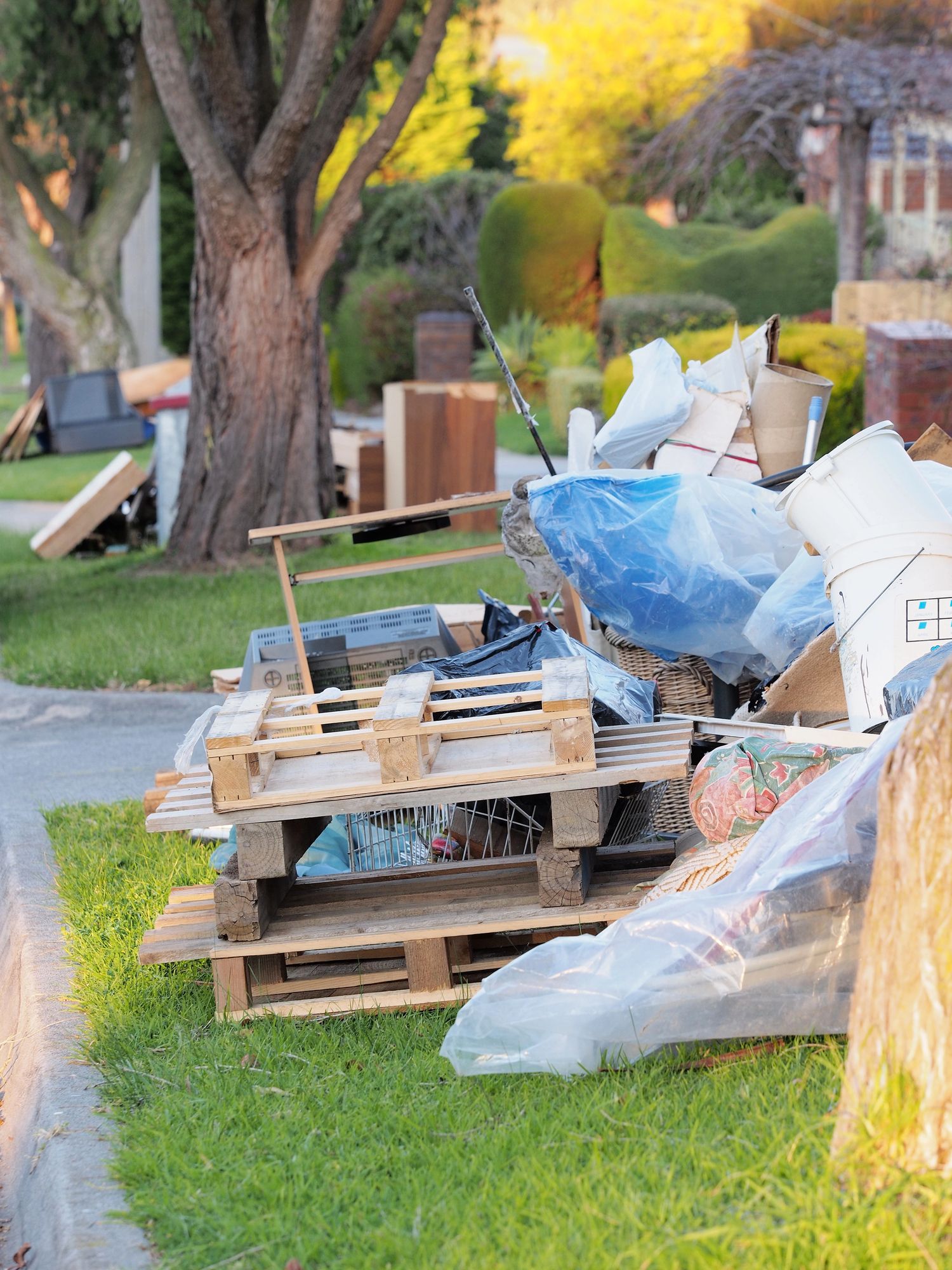 Pile of trash and wooden pallets on a grassy curb next to a sidewalk and trees.