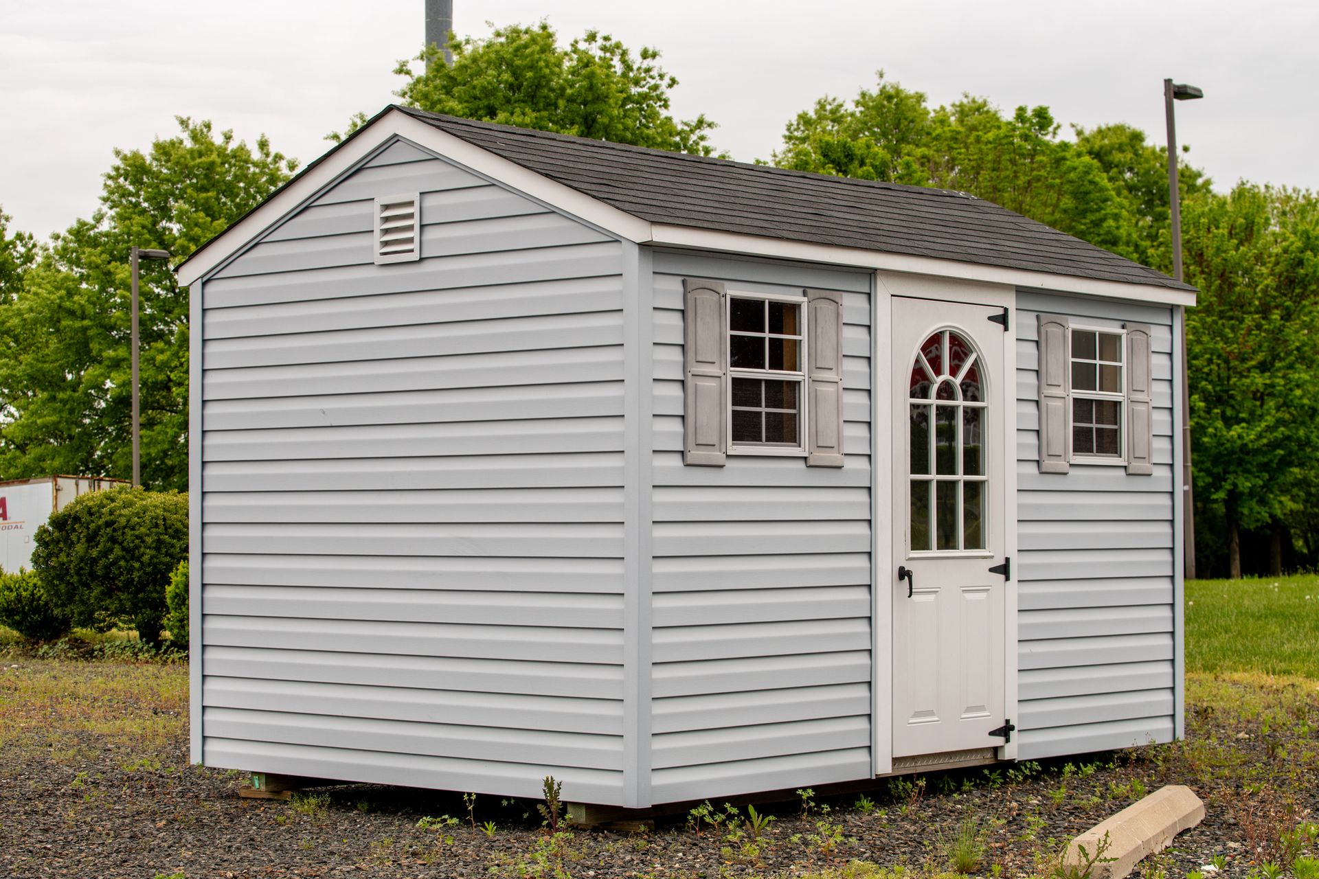 Light blue shed with a white door, windows, and shutters on a gravel lot.