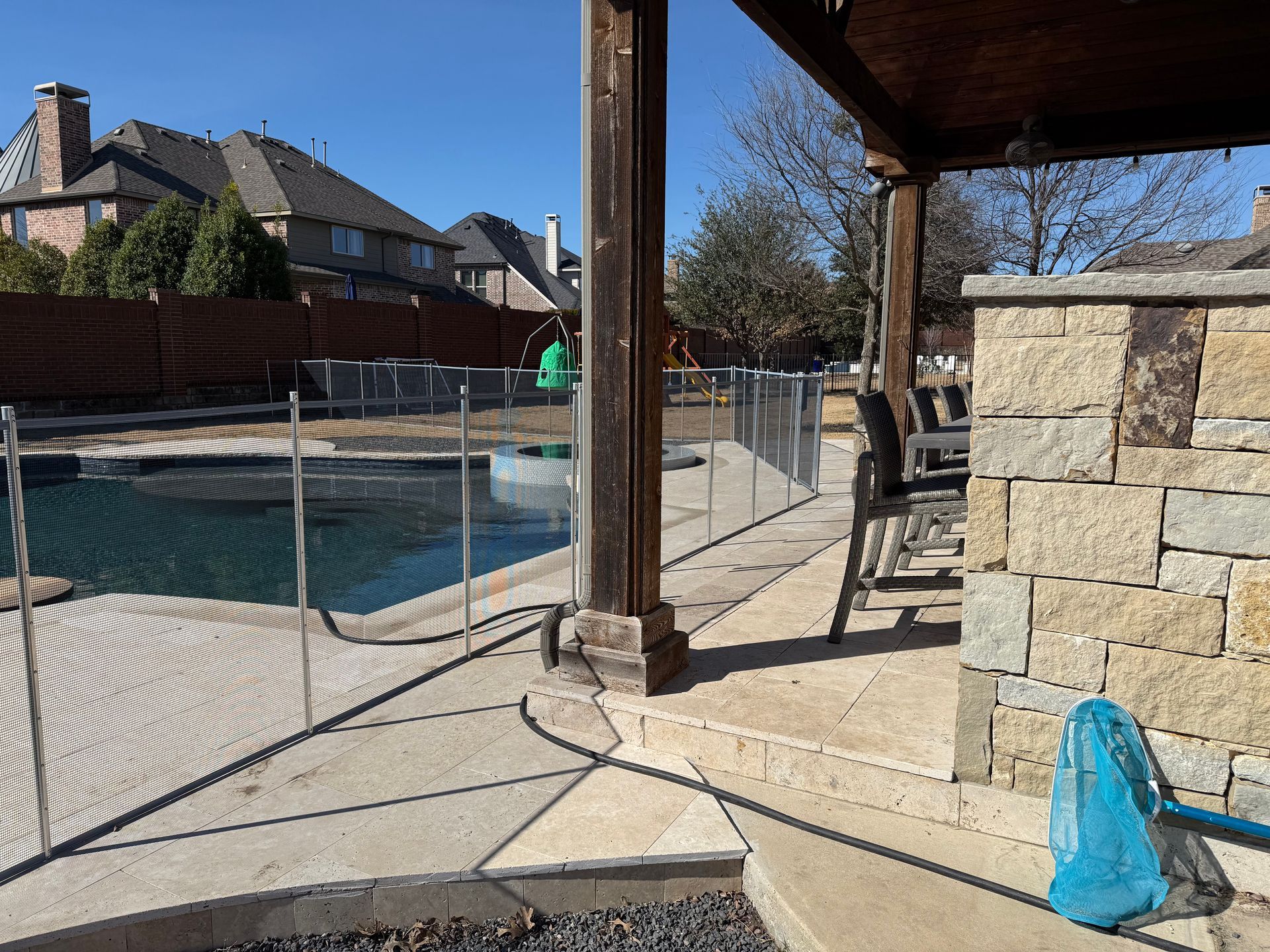 Covered patio beside a fenced backyard pool, with stone walls, columns, and a blue bag on the ground.