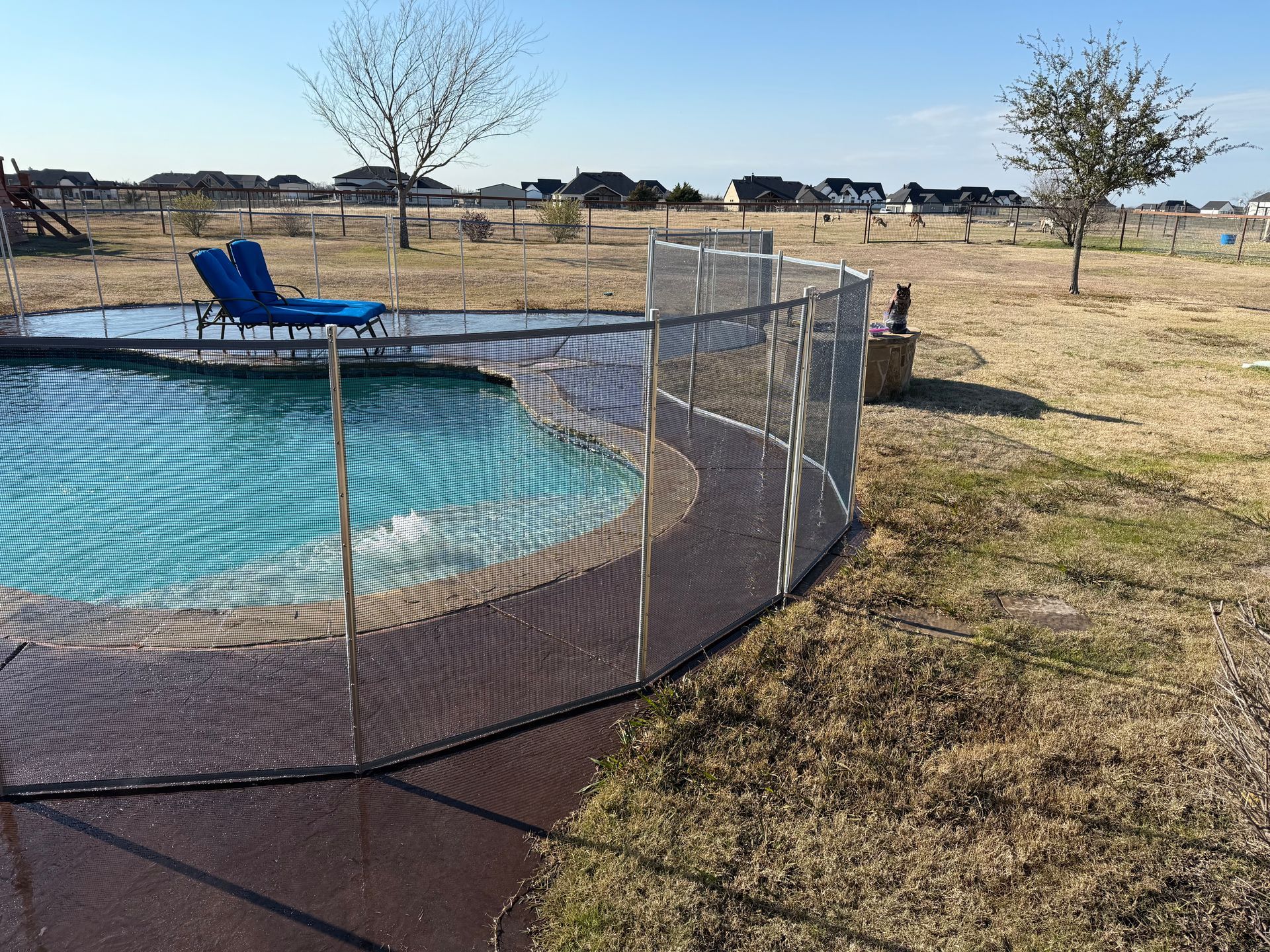 Mesh fence around an above-ground pool with a blue chair in a dry backyard.