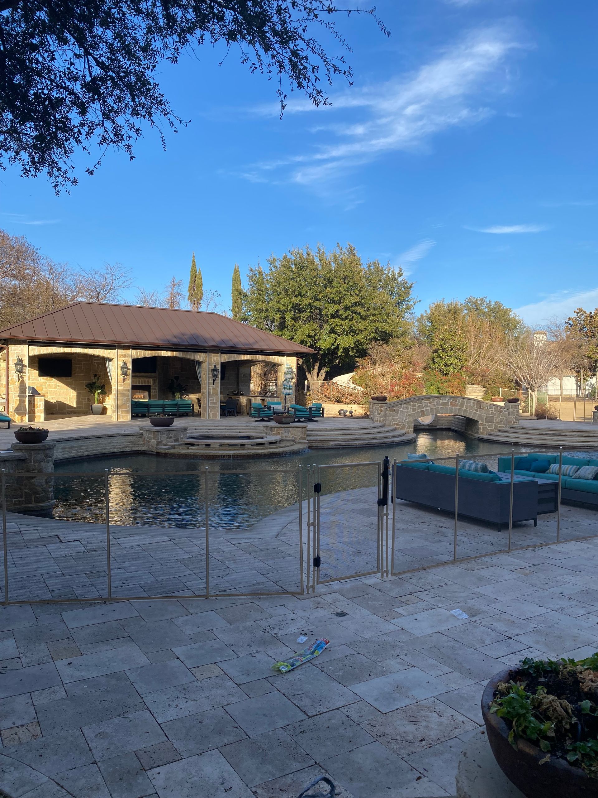 Backyard pool with stone patio, low fence, and a house under a clear blue sky