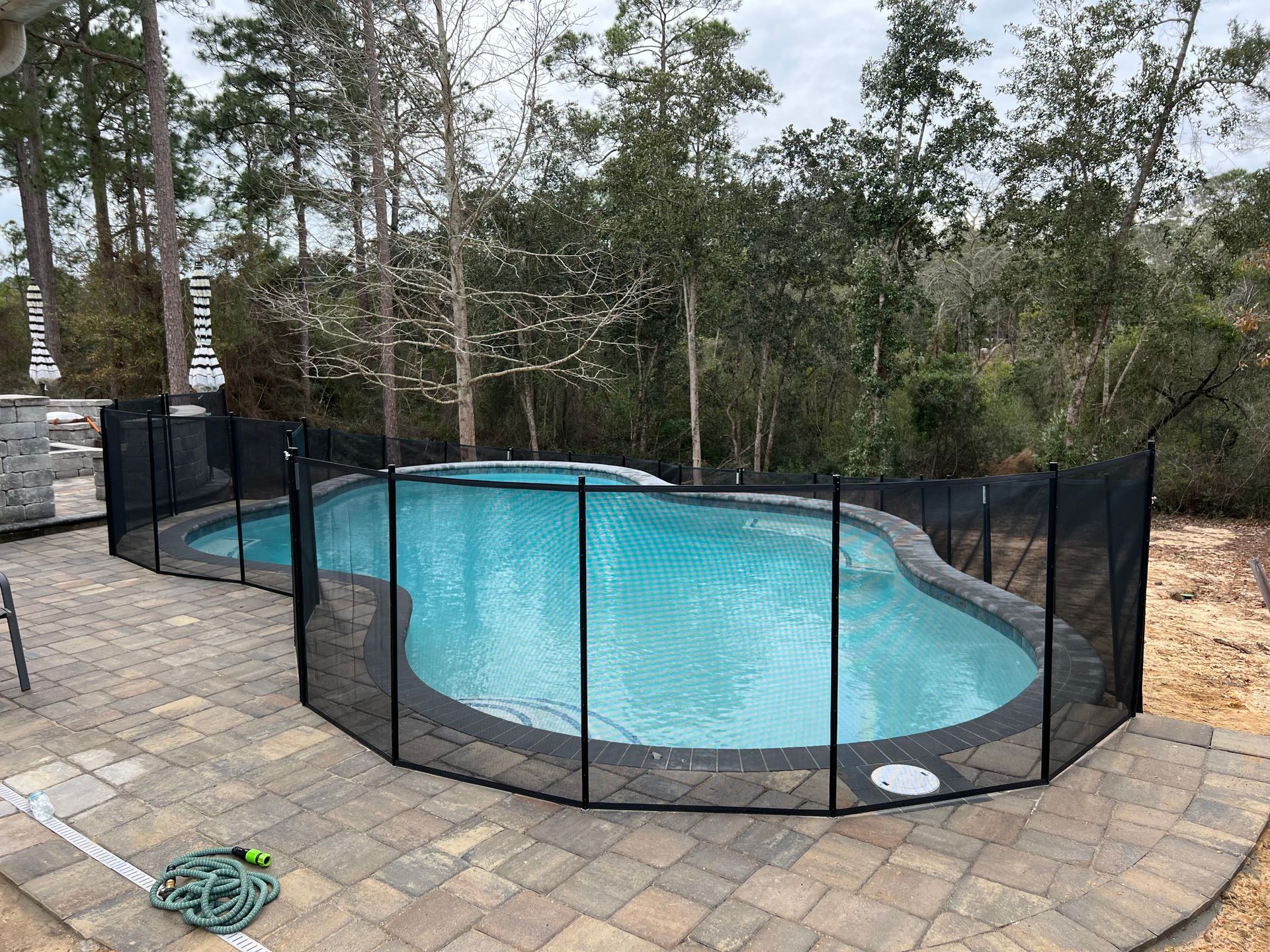 Outdoor pool with black safety fence on a stone patio, surrounded by trees