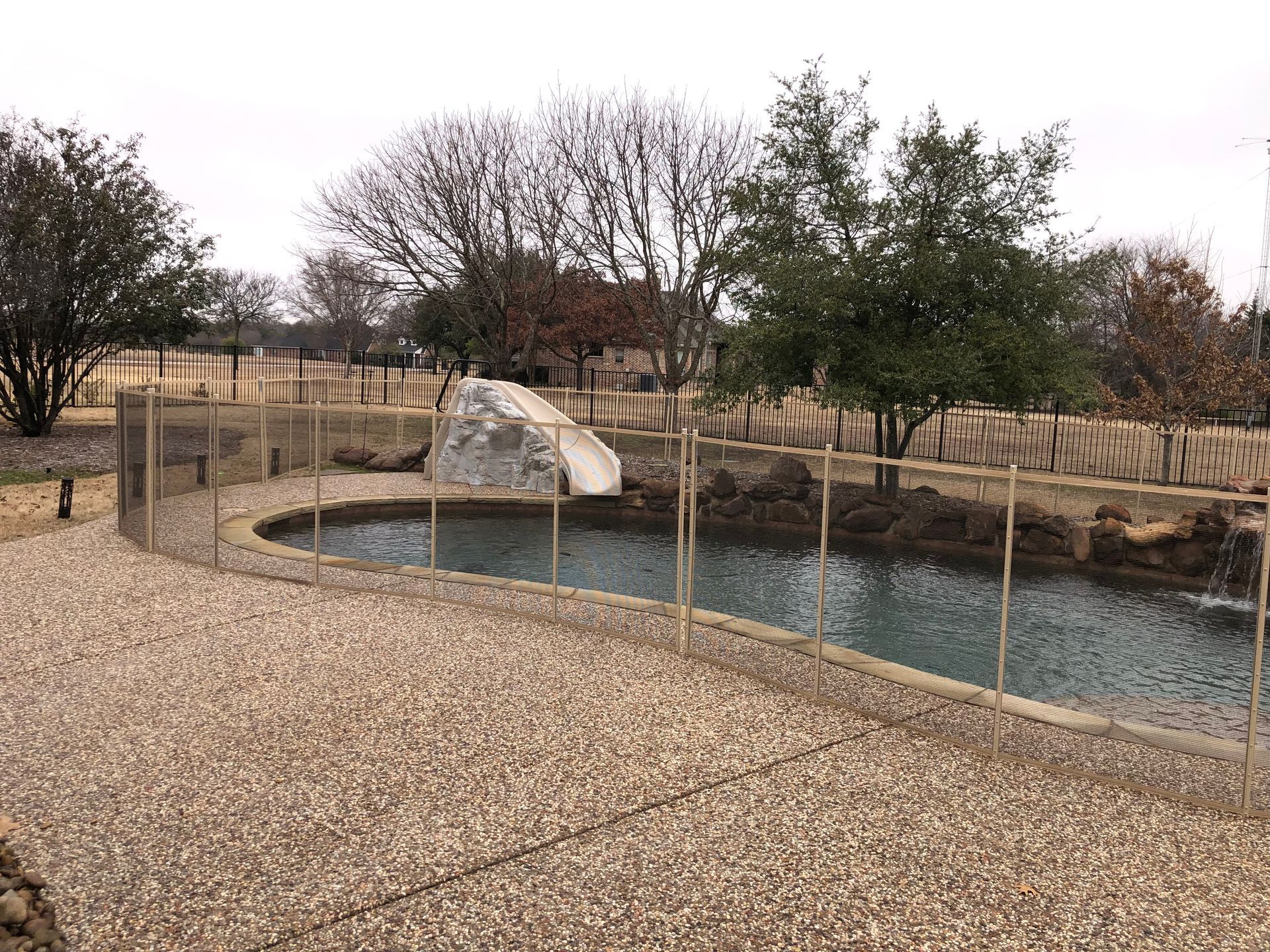 Small rocky pond in a fenced park with gravel path and trees under a cloudy sky
