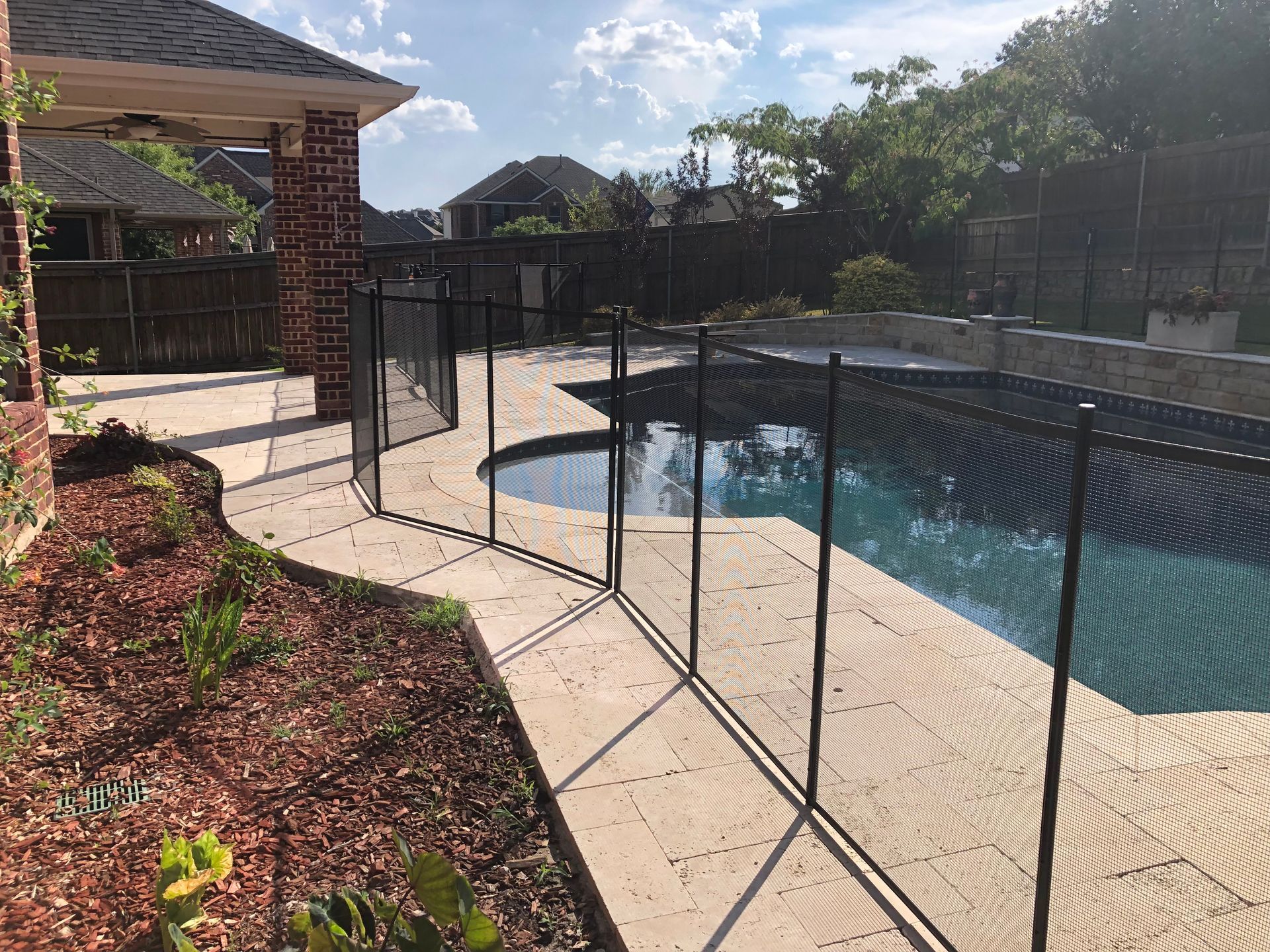 Backyard pool with black safety fence, stone patio, and landscaped garden in bright sunlight