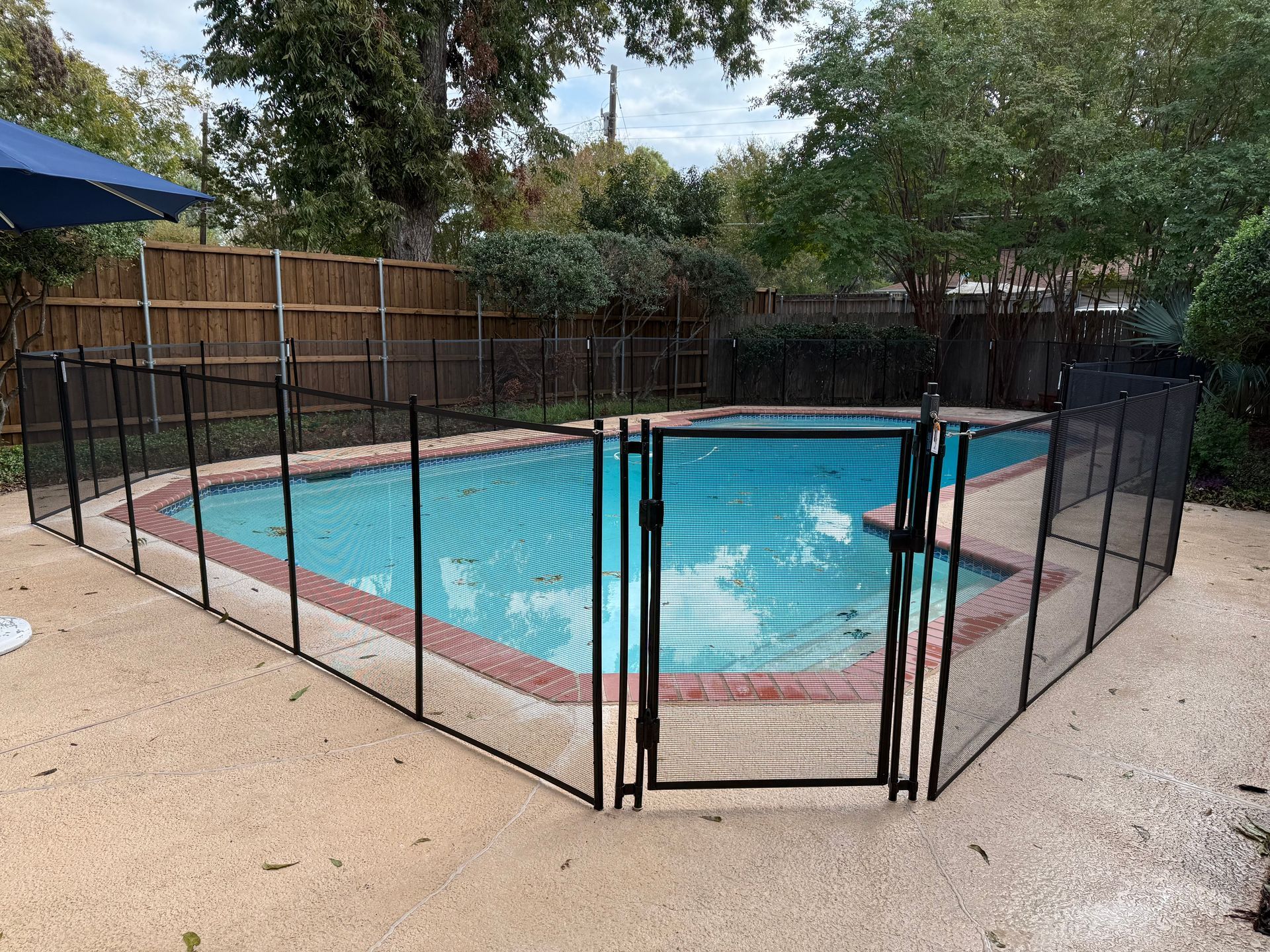 Backyard swimming pool enclosed by a black safety fence, with trees and patio around it