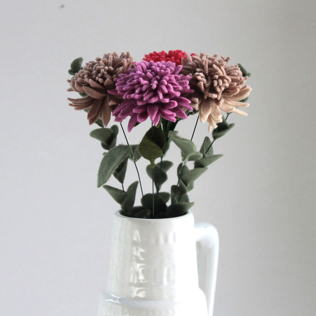 Boeket vilten asters in diverse kleuren, in een witte vaas tegen een witte achtergrond.
