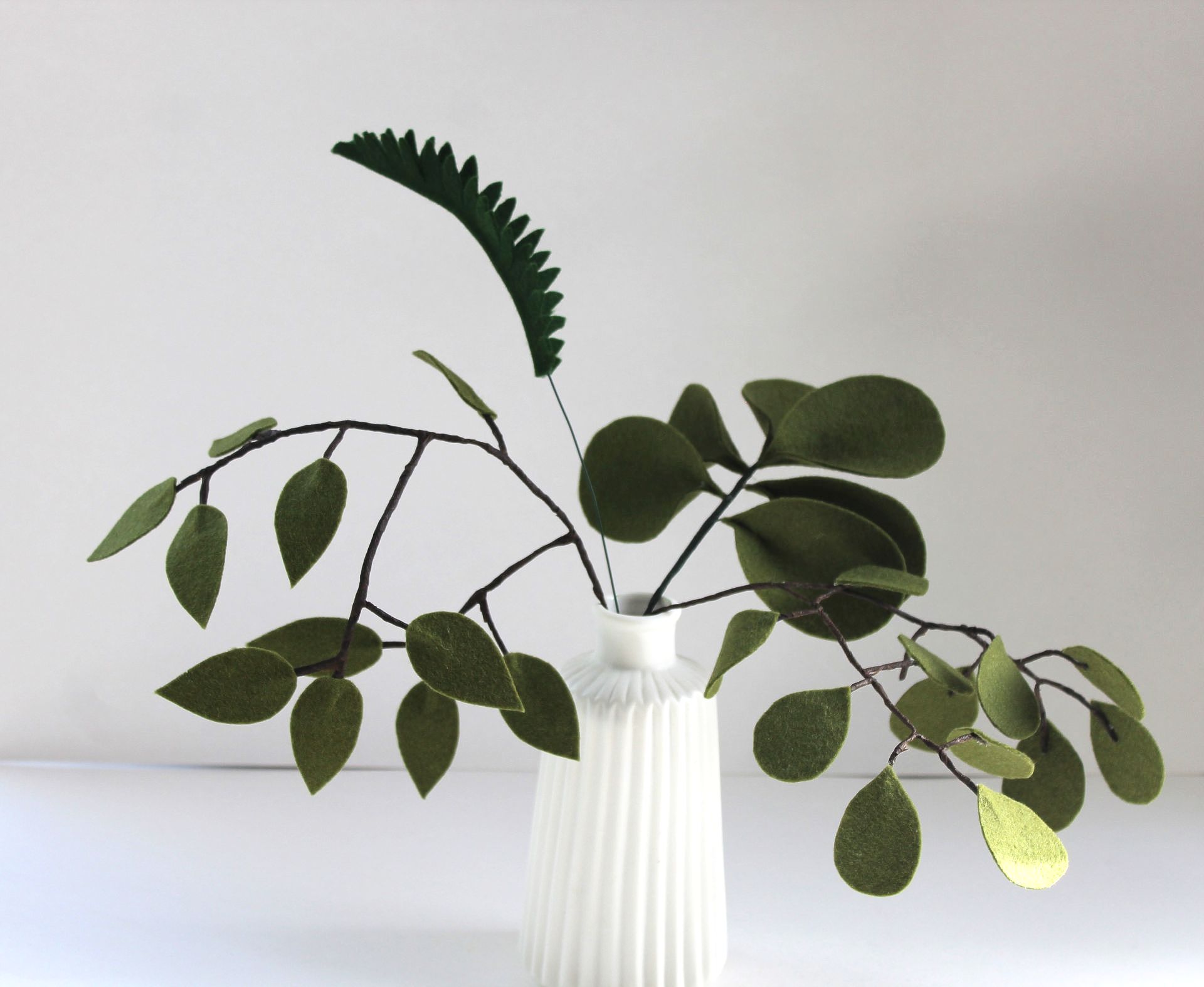 Viltbladeren in een witte vaas: eucalyptus- en varenachtige bladeren in donkergroene en saliegroene tinten.