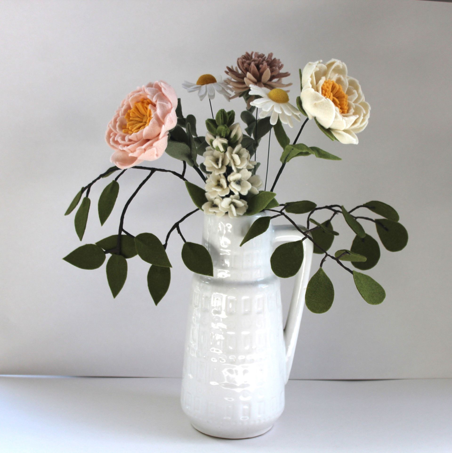 Witte vaas met vilten bloemen in roze, crème en wit, en groene bladeren.