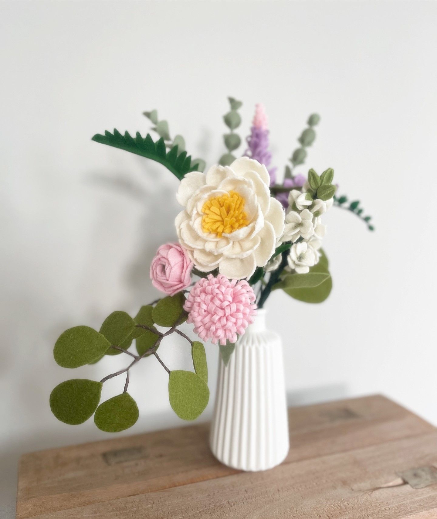 Bloemenarrangement van vilt in een witte vaas op een houten ondergrond. Met een witte pioenroos en roze en groene accenten.