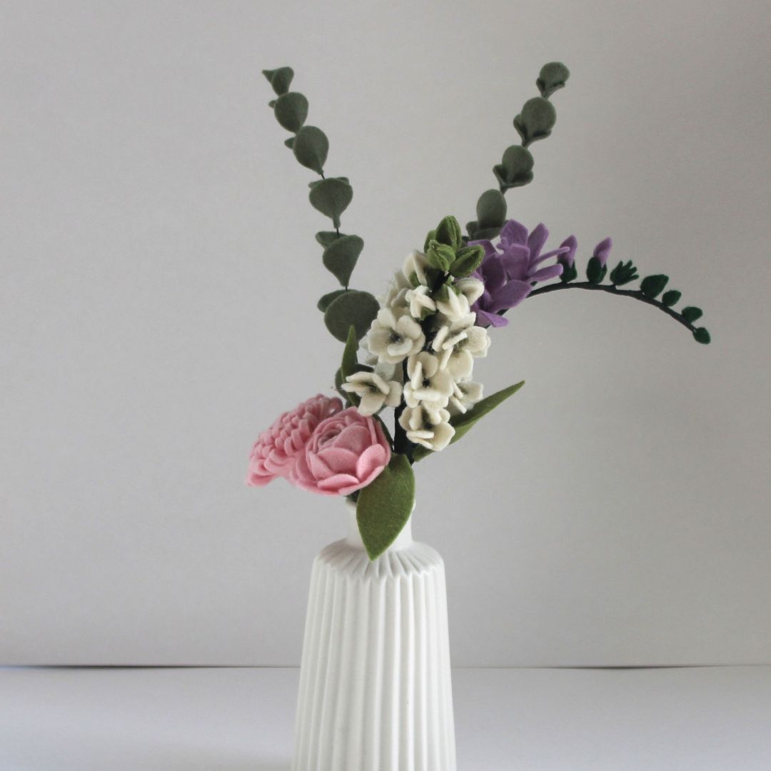 Bloemenarrangement van vilt in een witte vaas: roze pioenroos, witte stokroos, paarse fresia, groene eucalyptus.