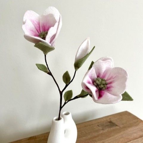 Vilten magnoliabloemen in een witte vaas op een houten ondergrond; roze en witte bloemblaadjes, groene bladeren.