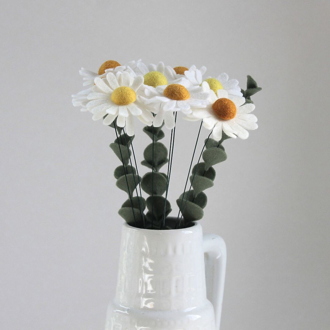 Witte madeliefjes met gele hartjes in een witte vaas, met groene eucalyptusbladeren.