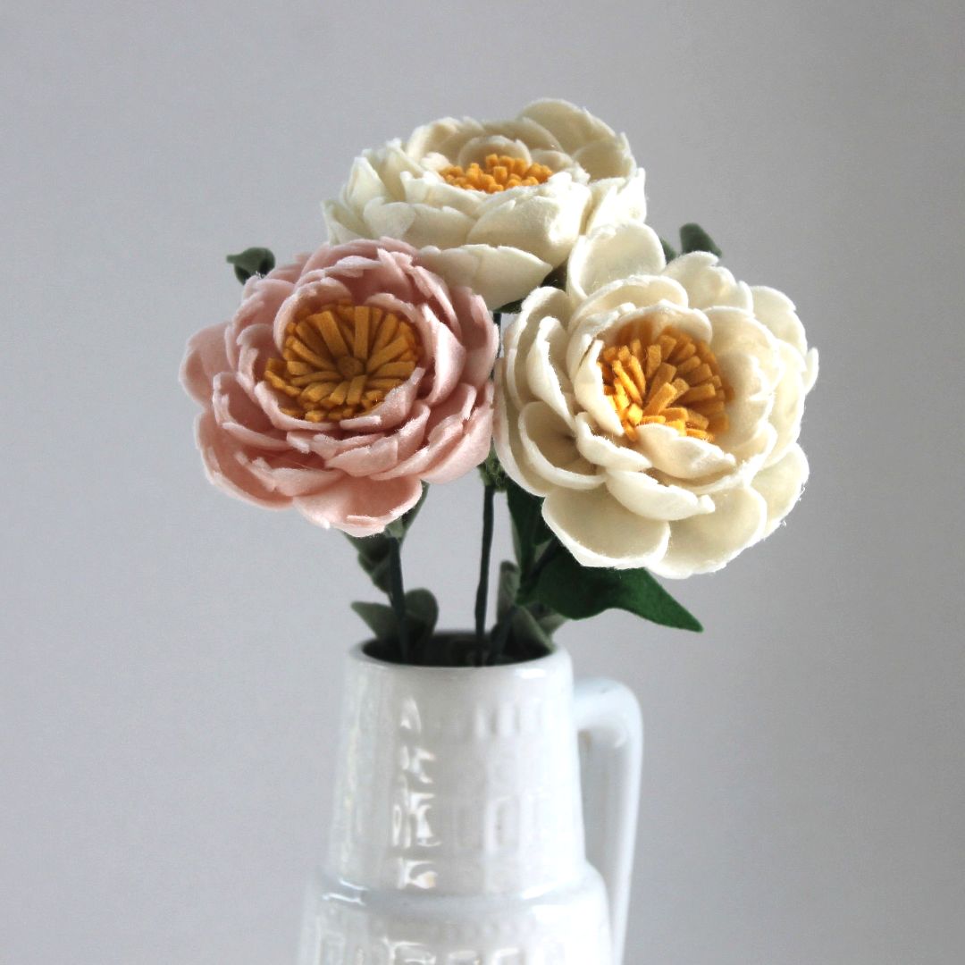 Drie vilten pioenrozen, roze en crèmekleurig, in een witte vaas.