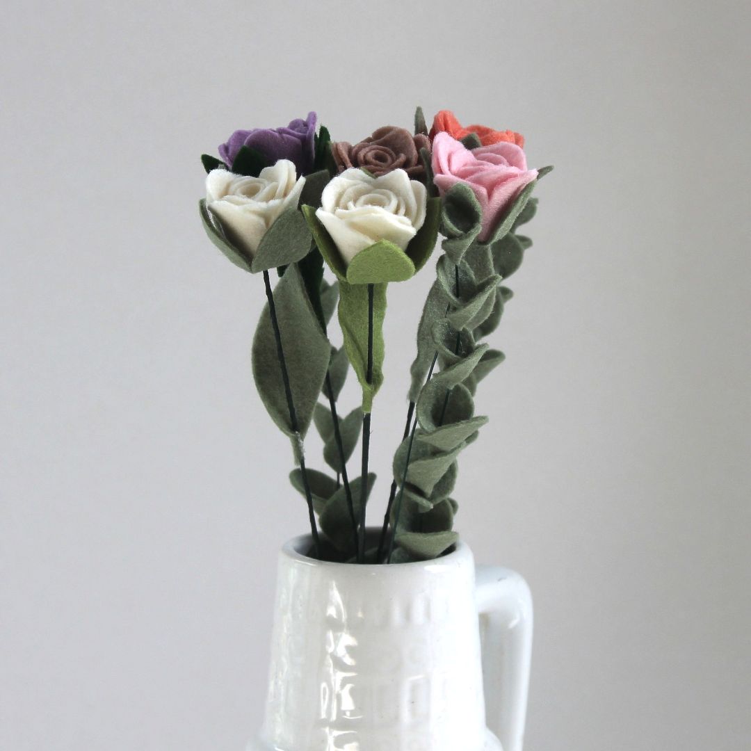Vilten rozen in diverse kleuren, geschikt in een witte vaas tegen een lichtgrijze achtergrond.