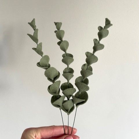 Een hand houdt drie groene, stoffen eucalyptusstengels vast tegen een witte achtergrond.