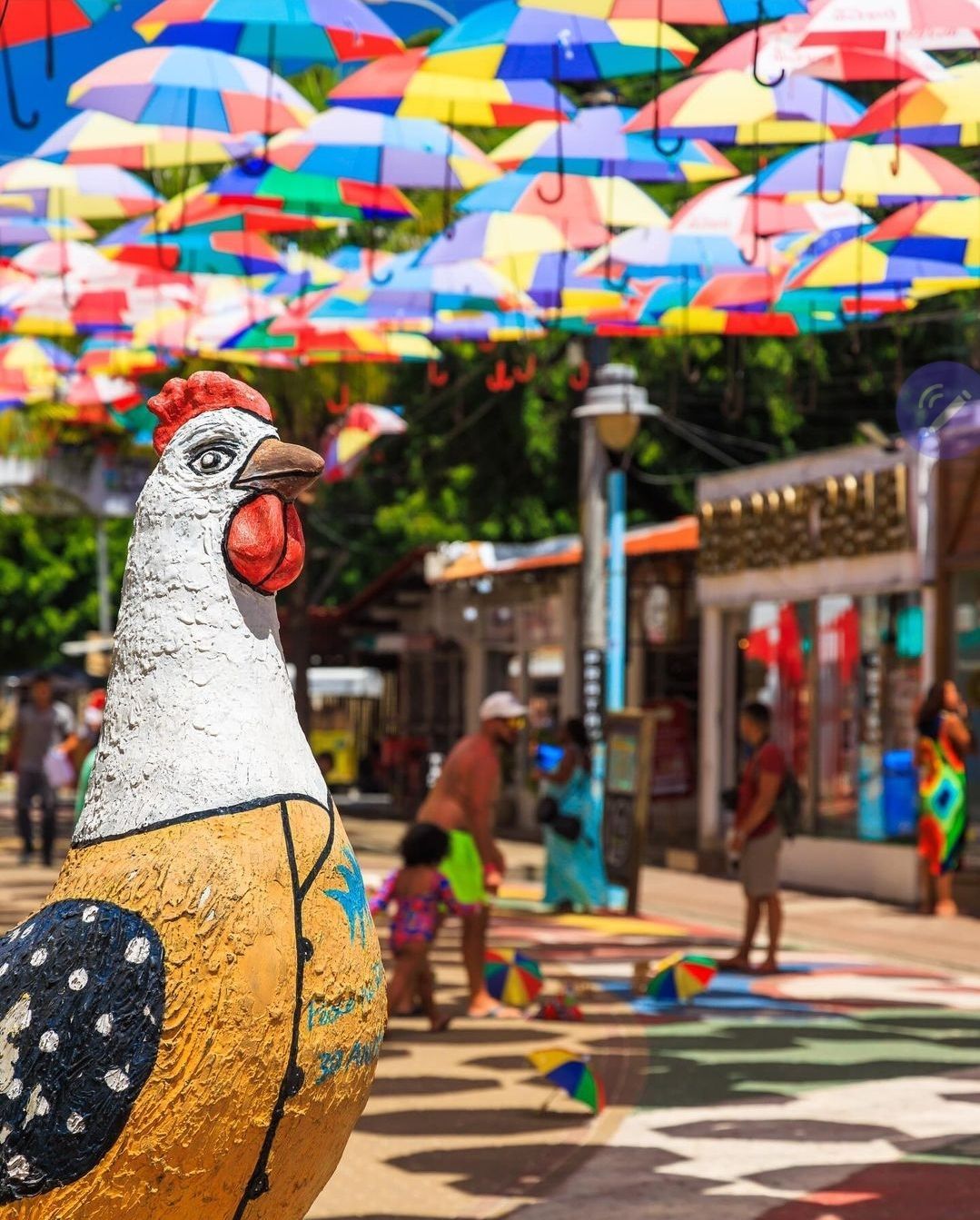 Uma estátua de uma galinha está em frente a uma fileira de guarda-chuvas coloridos.