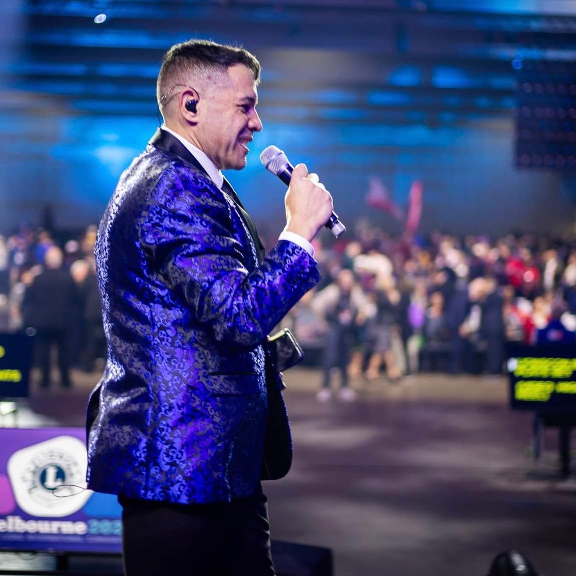 Man in a blue blazer speaking into a microphone on a stage before a blurred crowd