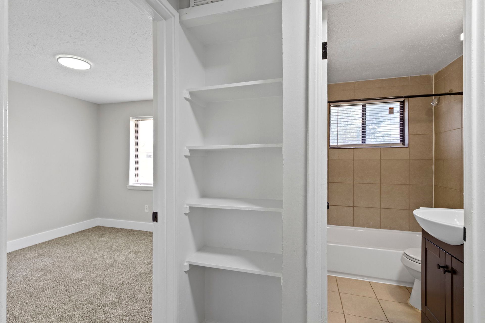 Hallway view of a room, storage shelves, and bathroom. Beige walls, beige tiled shower.