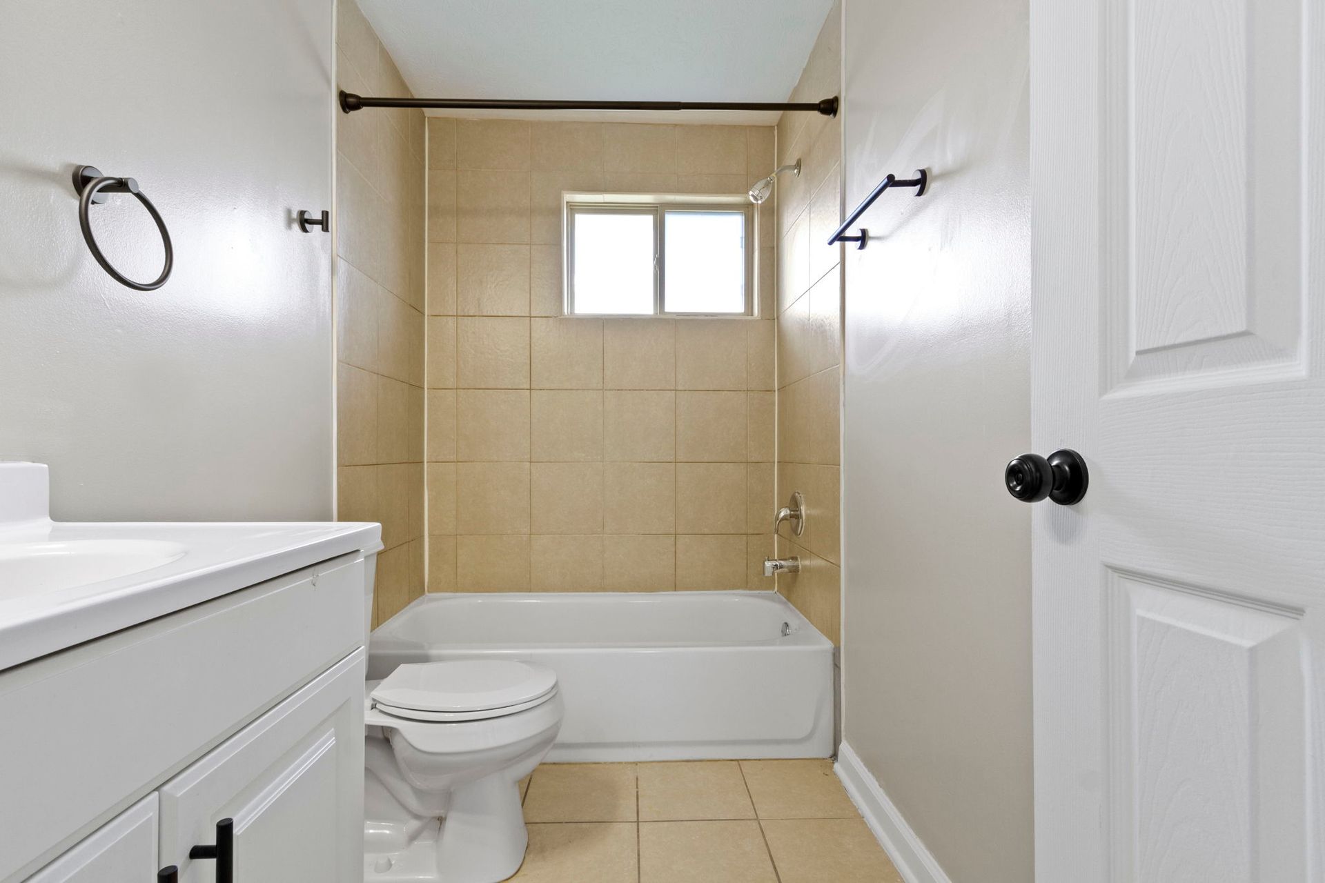 Bathroom with a white tub, toilet, and vanity, and beige tile. Black fixtures on gray walls.
