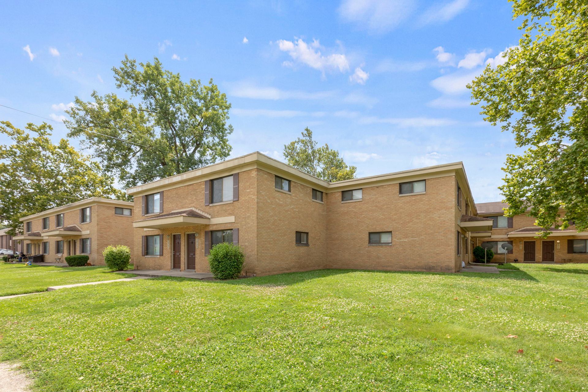 Brown brick apartment buildings with green lawn and trees under a blue sky.