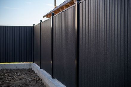 Tall black corrugated metal fence with concrete base along a dirt yard under a clear sky