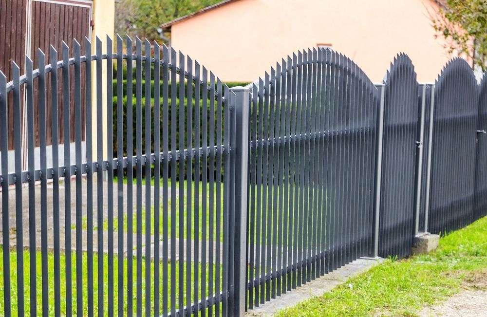 Black metal picket fence with arched gates along a grassy sidewalk beside a house