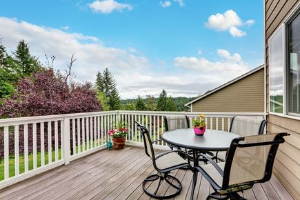 Deck with round table and chairs, potted flowers, white railing, and a scenic backyard view under a blue sky