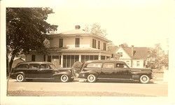 Sepia-toned vintage house with two classic cars parked out front under trees