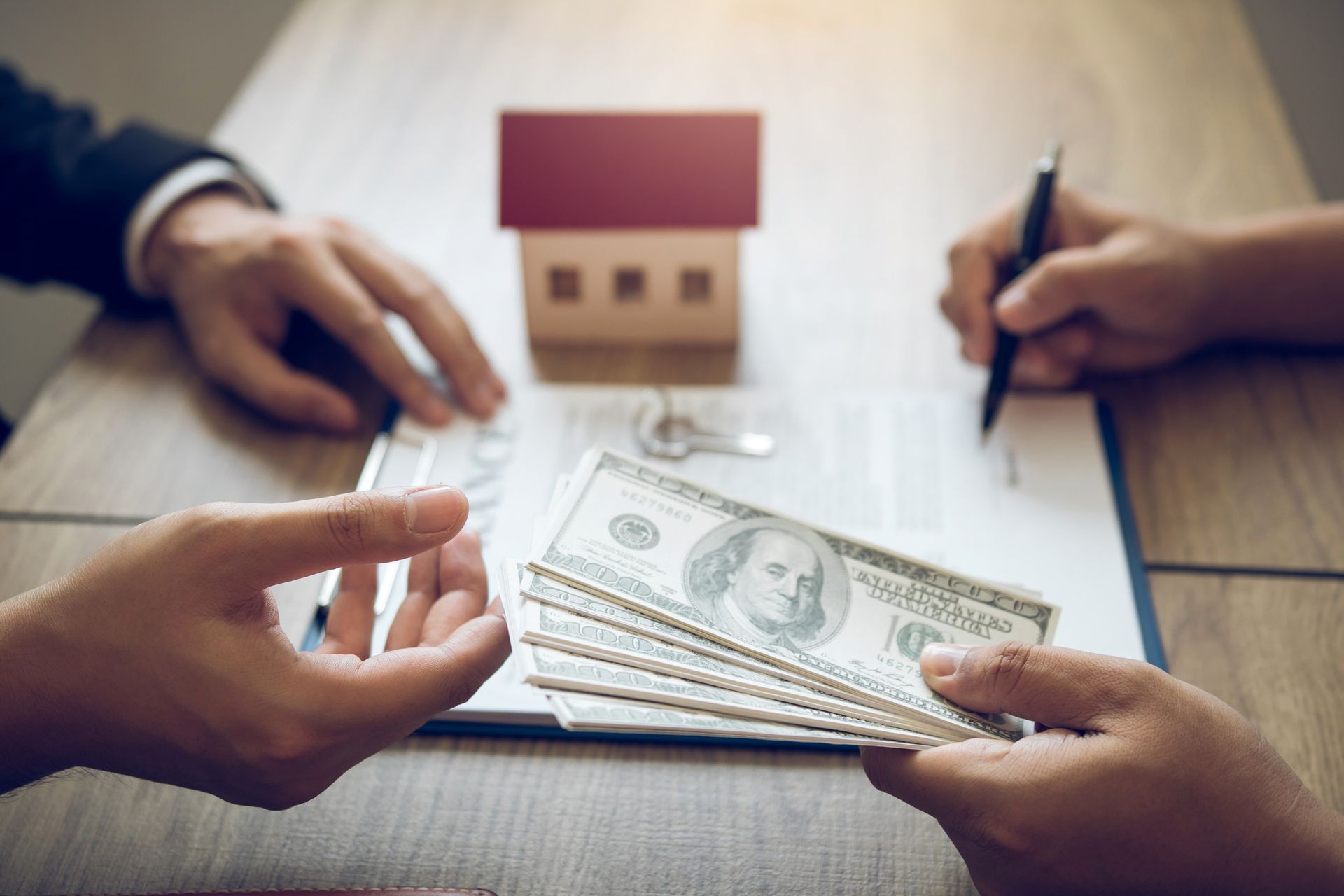 Hands exchanging cash over a contract and keys, with a model house on a desk.
